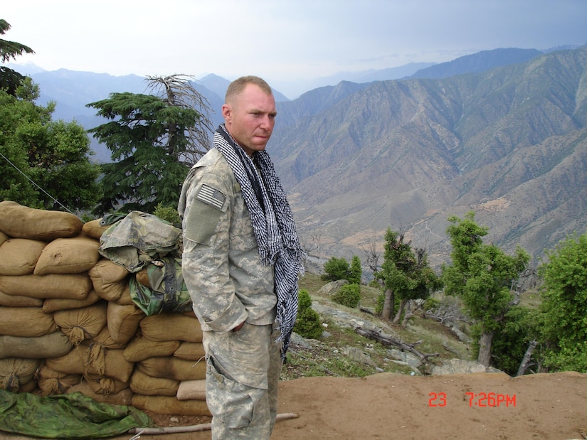 A man in a camouflage military uniform looks into the distance as he stands on the side of a mountain. There are trees and a mountain range in the background.