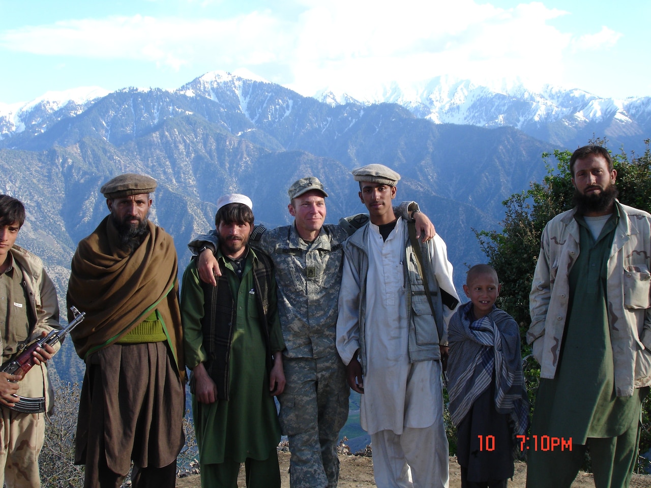 A man wearing a military camouflage uniform, center, five other men and a child pose for a photo with mountains in the background.