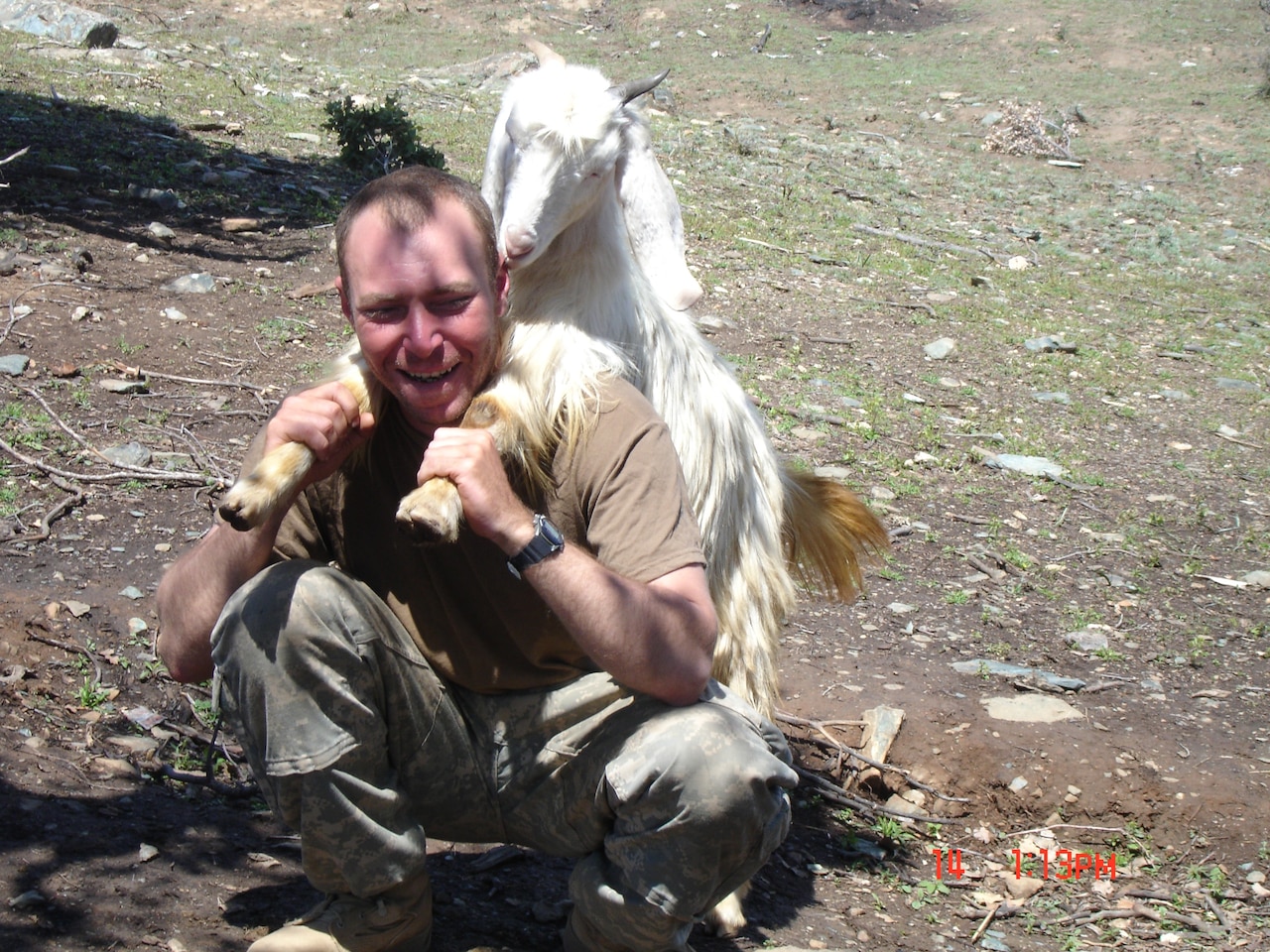 A smiling man who is kneeling down holds a goat's front legs over his shoulders.