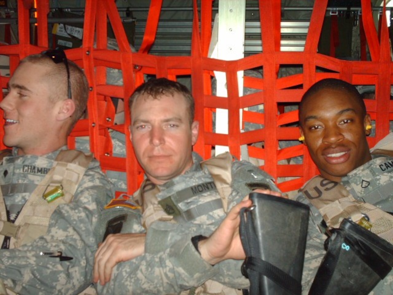 Three men wearing military camouflage uniforms lie down in a cargo aircraft bay holding long guns and posing for a photo.