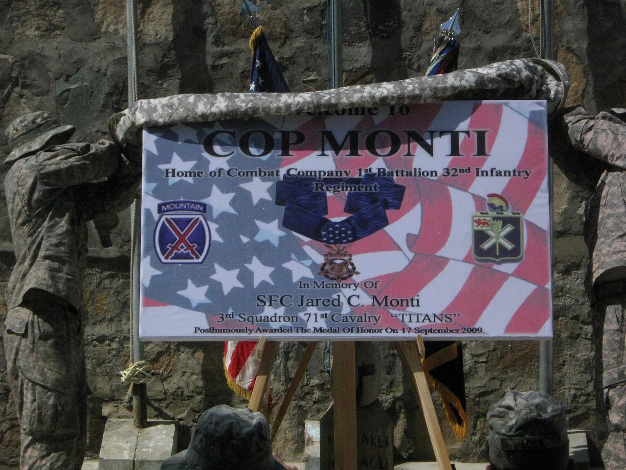 Two men in camouflage military uniforms pull a blanket off a sign to unveil it.