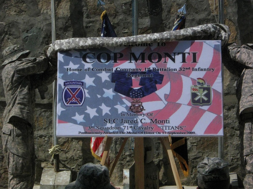 Two men in camouflage military uniforms pull a blanket off a sign to unveil it.