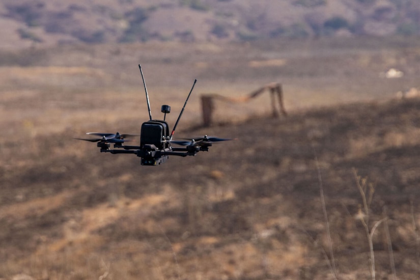 A small, black drone hovers over a desert landscape.