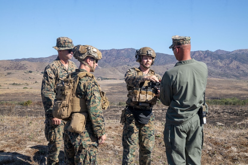 Four people wearing military uniforms stand outside in a desert environment with mountains in the background. Two look on as the person in the middle hands a small, black drone to the person standing the furthest right.