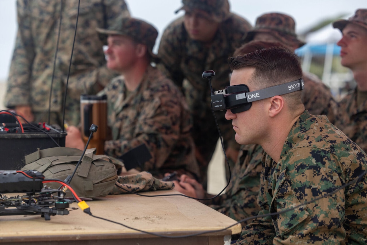 A person wearing a camouflage military uniform wears a headset to monitor an unmanned aerial system that is not pictured. People wearing similar attire are in the background, also looking at something not pictured.