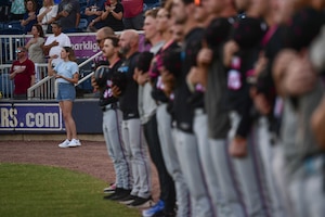 A female sings into a microphone next to a line of baseball players.