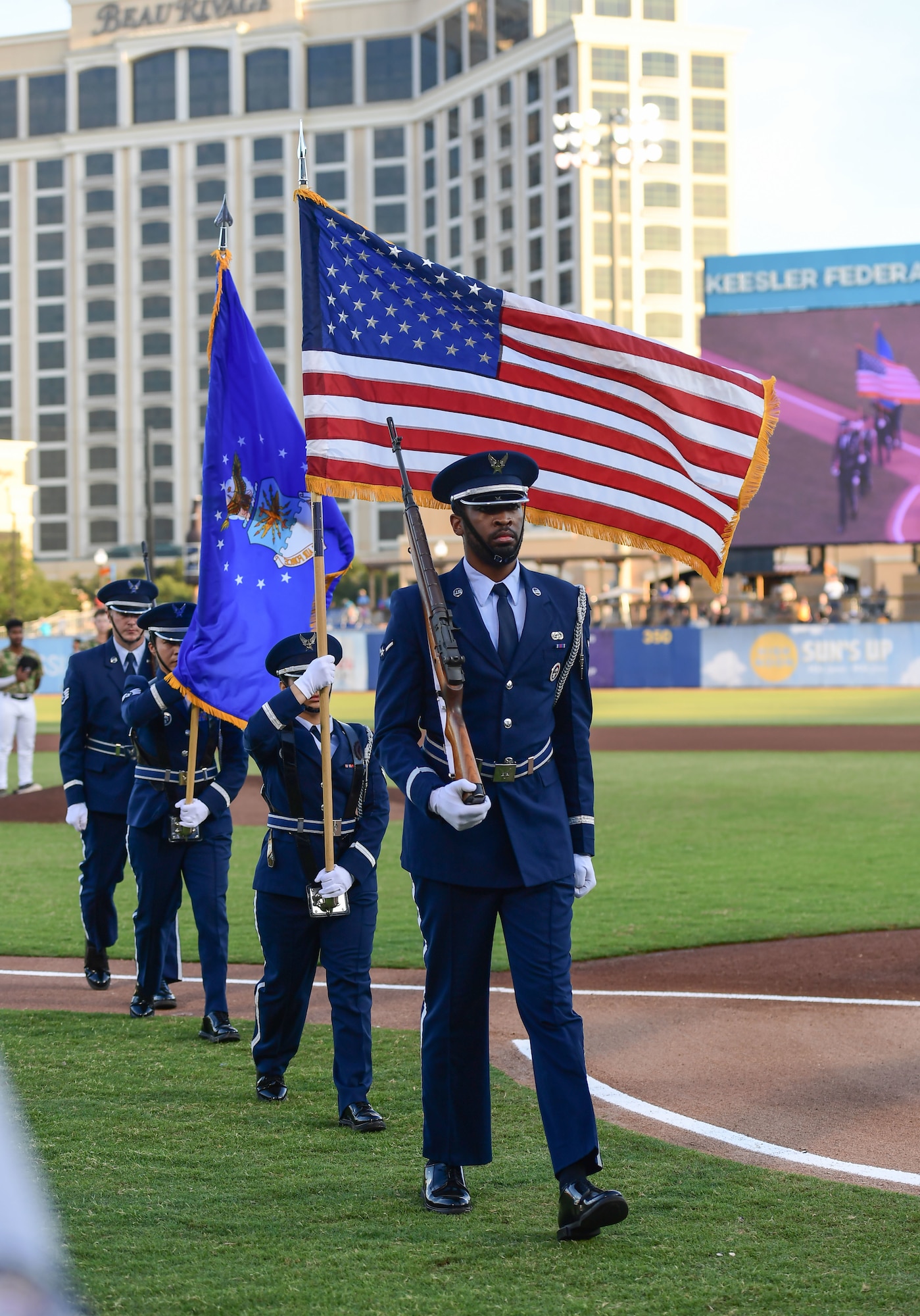 People in military uniforms carry flags while marching off a baseball field.