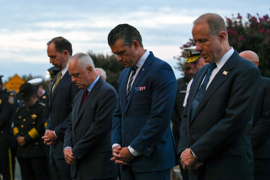 Four men in suits bow their heads and cross their arms respectfully in front.