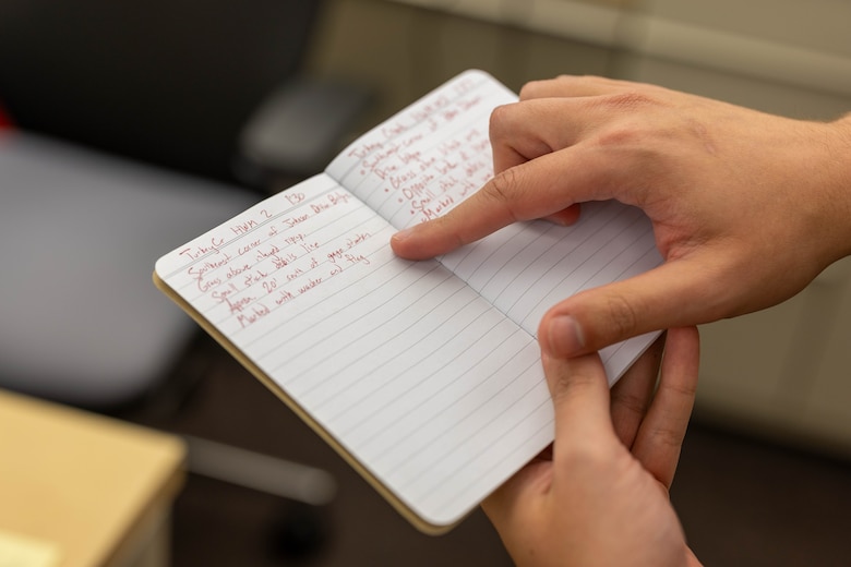 Two hands hold a small notebook with lined paper and red ink notes written on the inside.