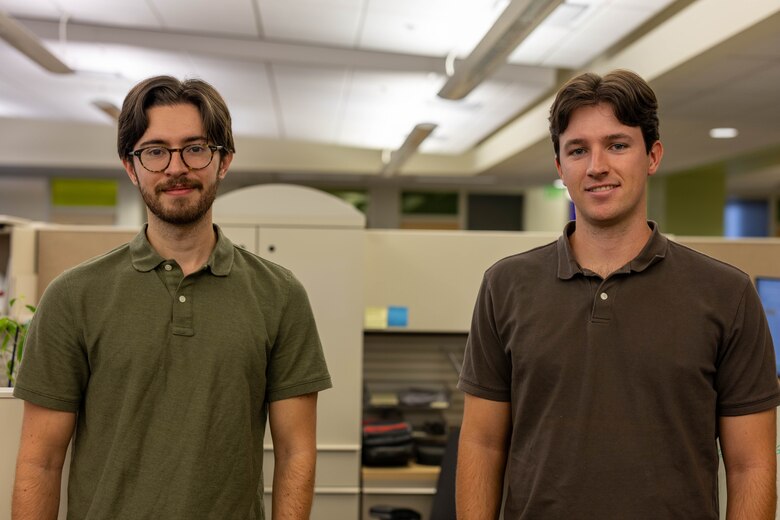 Two men, one with a green shirt and glasses and one with a brown shirt, stand and smile at the camera.