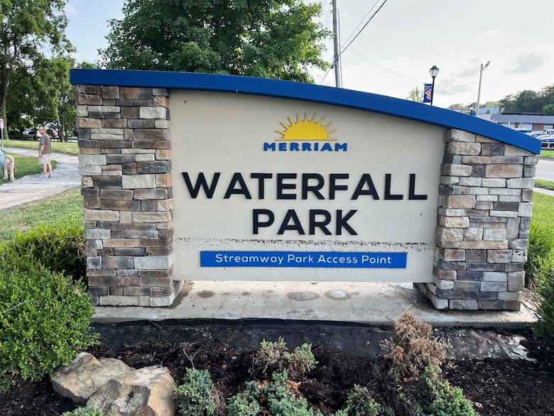 A stone and concrete park sign with brown mulch and green plants in the foreground and trees and a grey sky in the background.