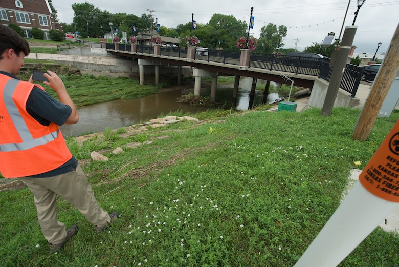 A man in an orange safety vest takes a photo of a line of sticks and debris on a hill with green grass with a creek to the left and a grey concrete bridge in the background.