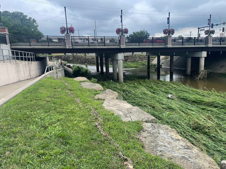 A line of sticks and debris sits on a green hill with a creek on the right side and a grey concrete bridge in the background.
