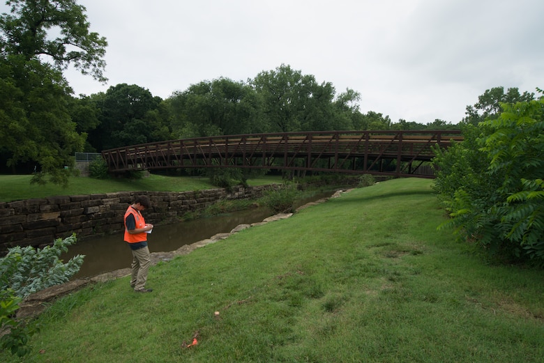 A man in an orange safety vest and khaki pants stands on a hill and writes in a notebook with a creek and a brown bridge, green grass and trees, and a grey sky in the background.
