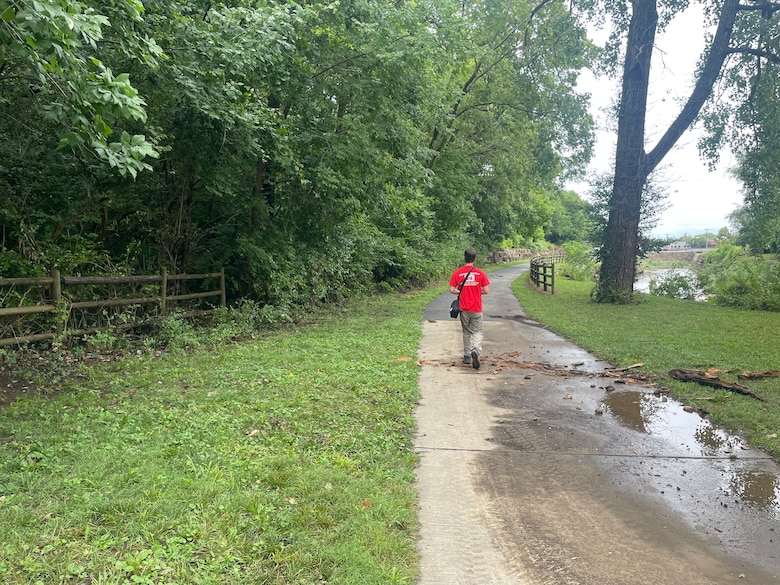 A man in a red shirt and khaki pants walks on a concrete trail with grass and trees on both sides.