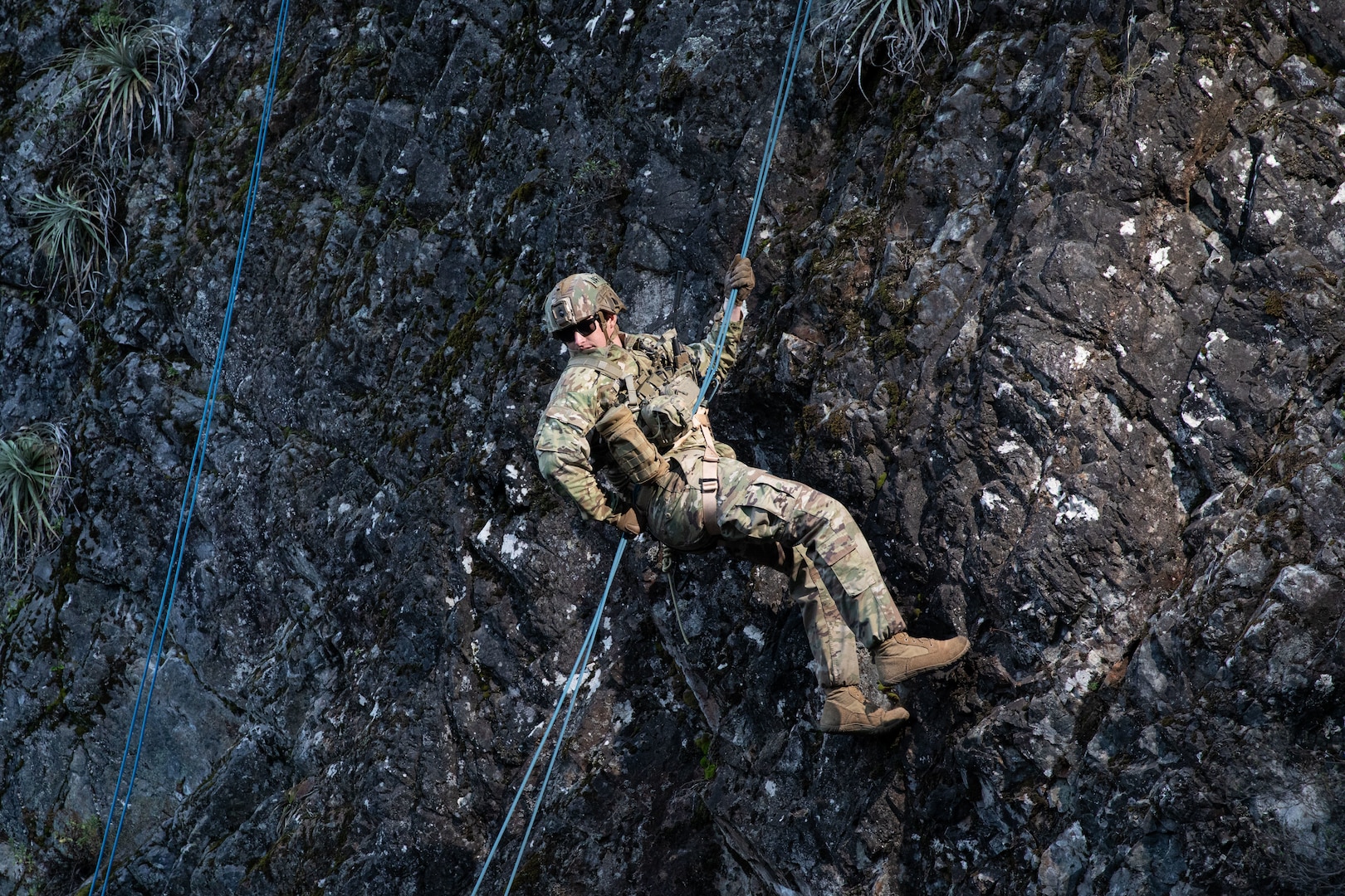 A man in a camouflage uniform and helmet rappels down a mountainside.