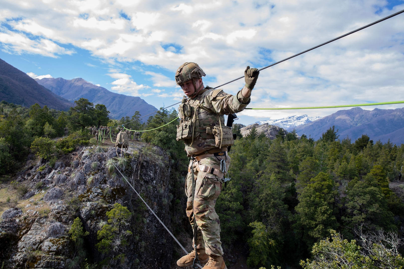 A man in a camouflage uniform and helmet crosses a two-rope bridge in the mountains.