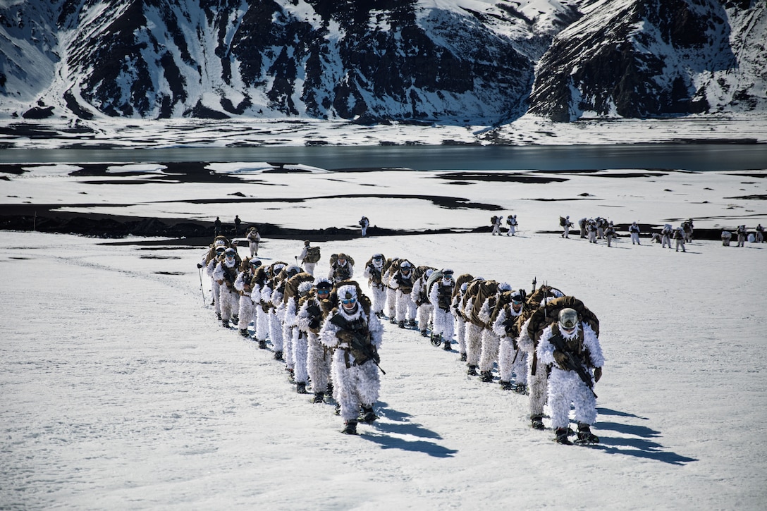 Two lines of personnel in white cold weather suits with large backpacks, snow shoes and trekking poles walk in a snowy, mountainous region.