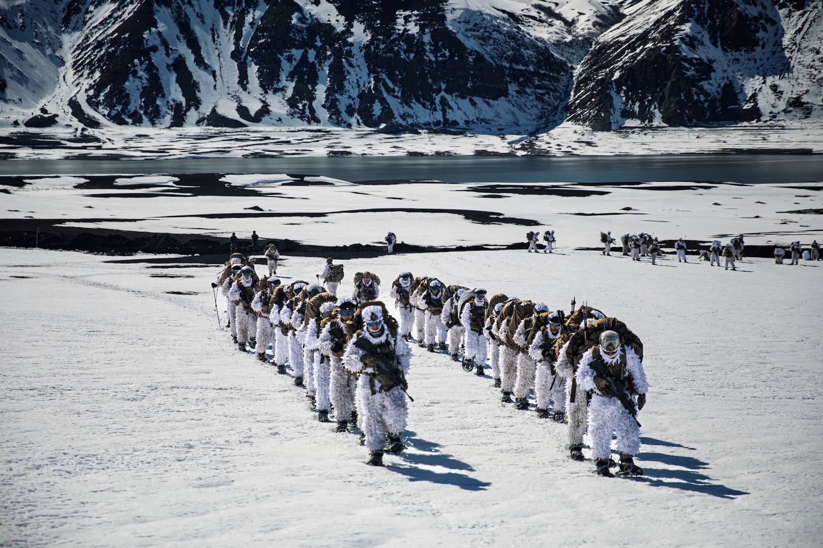 Two lines of personnel in white cold weather suits with large backpacks, snow shoes and trekking poles walk in a snowy, mountainous region.