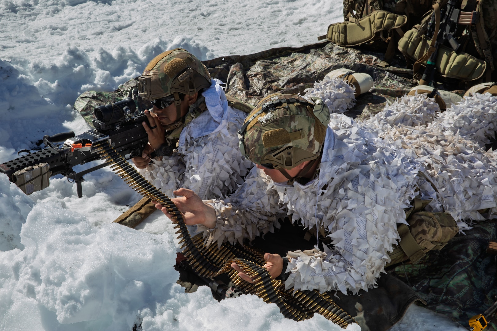 Men in while snow suits lay in the snow and shoot a rifle.