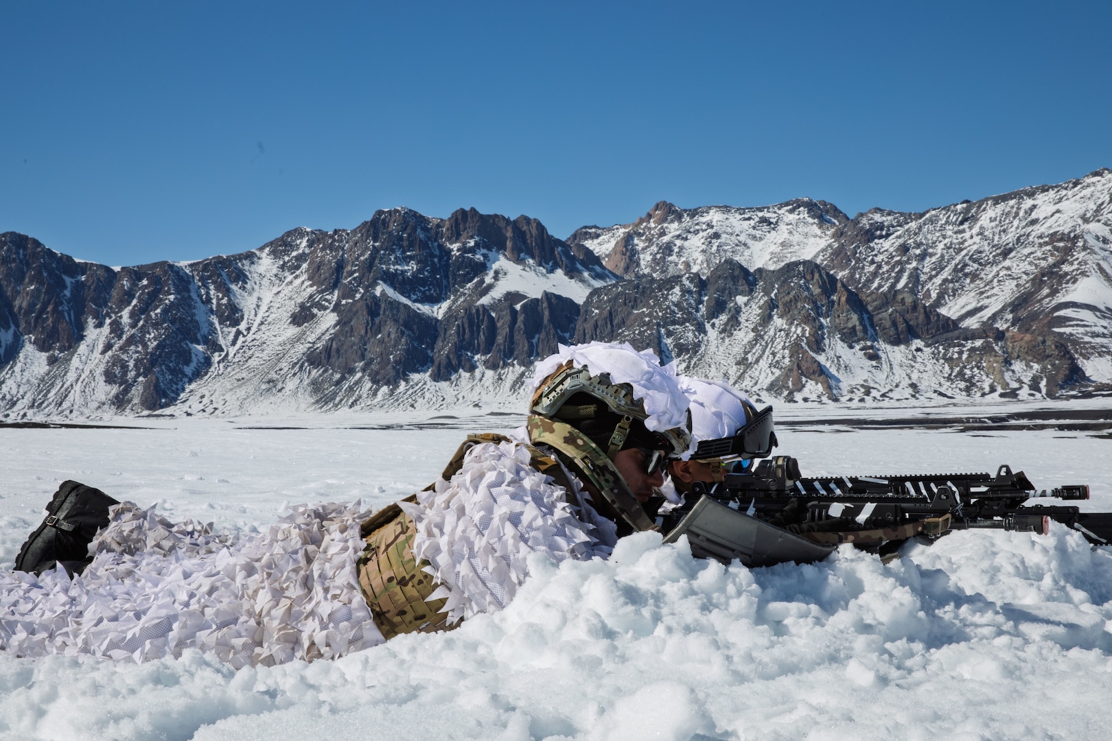 Two men in white cold weather suits lay in the snow and point rifles.