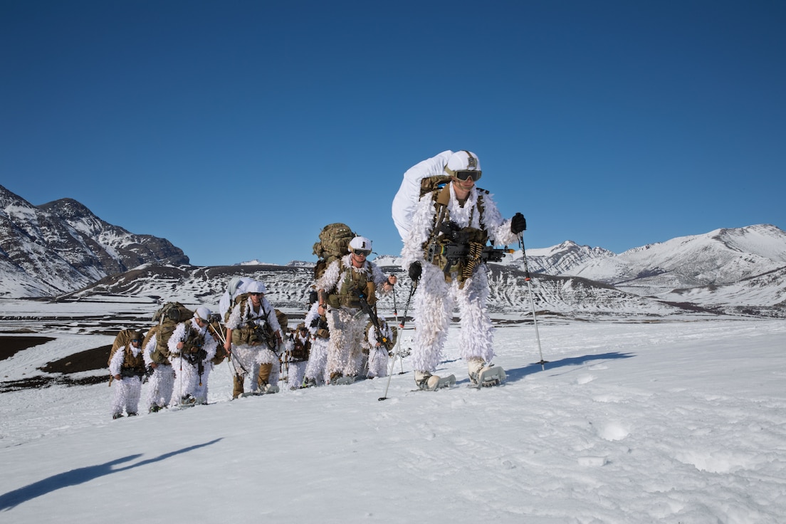 A line of personnel in white cold weather suits with large backpacks, snow shoes and trekking poles walk in a snowy, mountainous region.