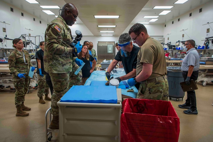 A man wearing a face shield, cuts open a boot as several people dressed in camouflage military uniforms or civilian attire watch in a laboratory setting. There is one man standing at an elevated position holding a camera.