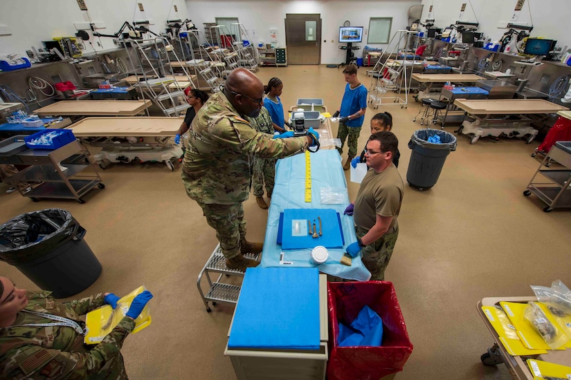 A man dressed in a camouflage military uniform and standing on a foot stool is taking a photograph of human remains. There are several others in similar attire watching or taking notes.