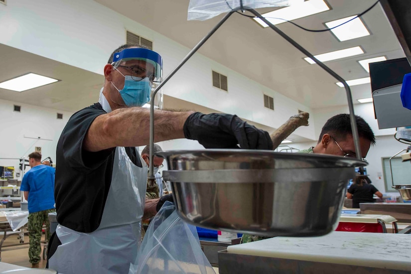A man wearing a protective face shield and a plastic apron places human remains on to a scale. There is a man in the background also wearing a protective face shield taking notes and a man wearing a camouflage military uniform.