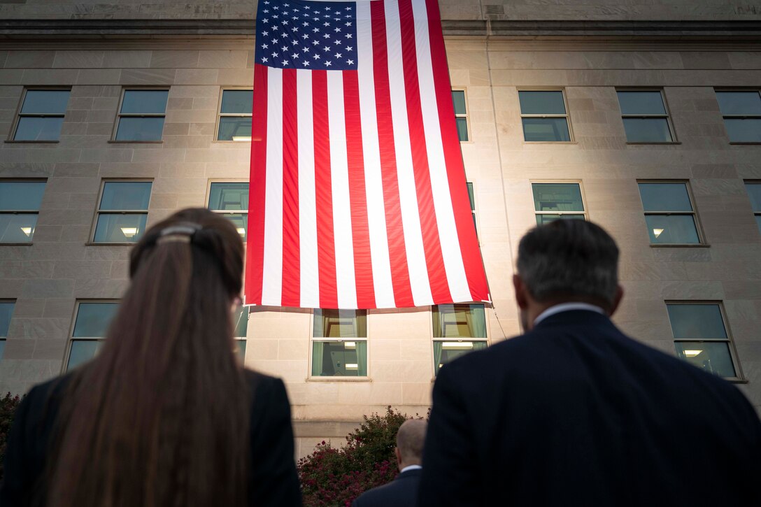 Two people, shown from behind look toward a large American flag hanging off the side of a building.
