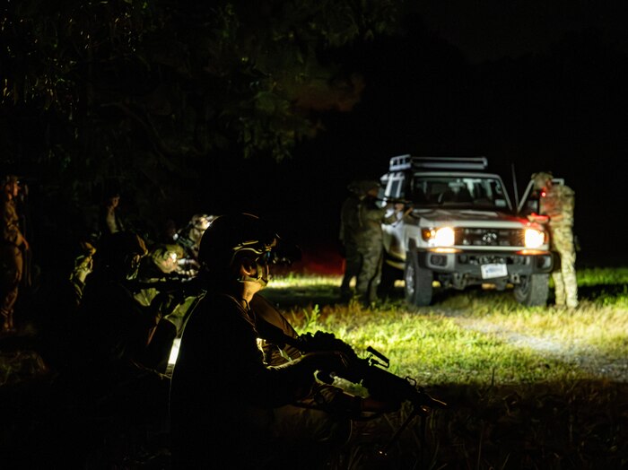 Two soldiers in camouflage uniforms stand beside a vehicle at night. The vehicle has its headlights on, and there are additional soldiers in similar attire kneeling in the foreground. The area in the foreground is dark, and the soldiers are holding rifles.