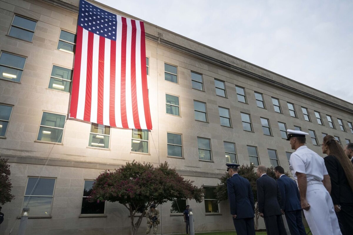 A large American flag hangs off the side of a large, five-story building at sunrise. Service members in military attire face the flag and stand at attention.