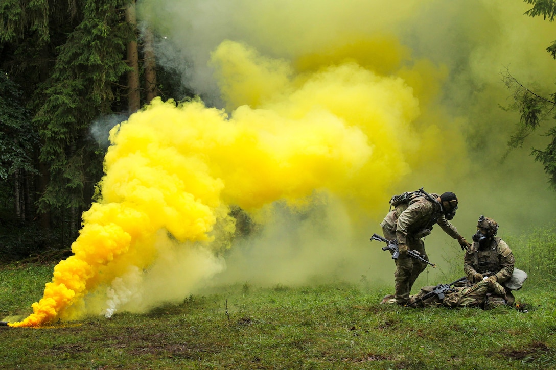 Two people wearing camouflage military uniforms and gas masks talk in a wooded, grassy environment, as a yellow plume of smoke drifts toward them.