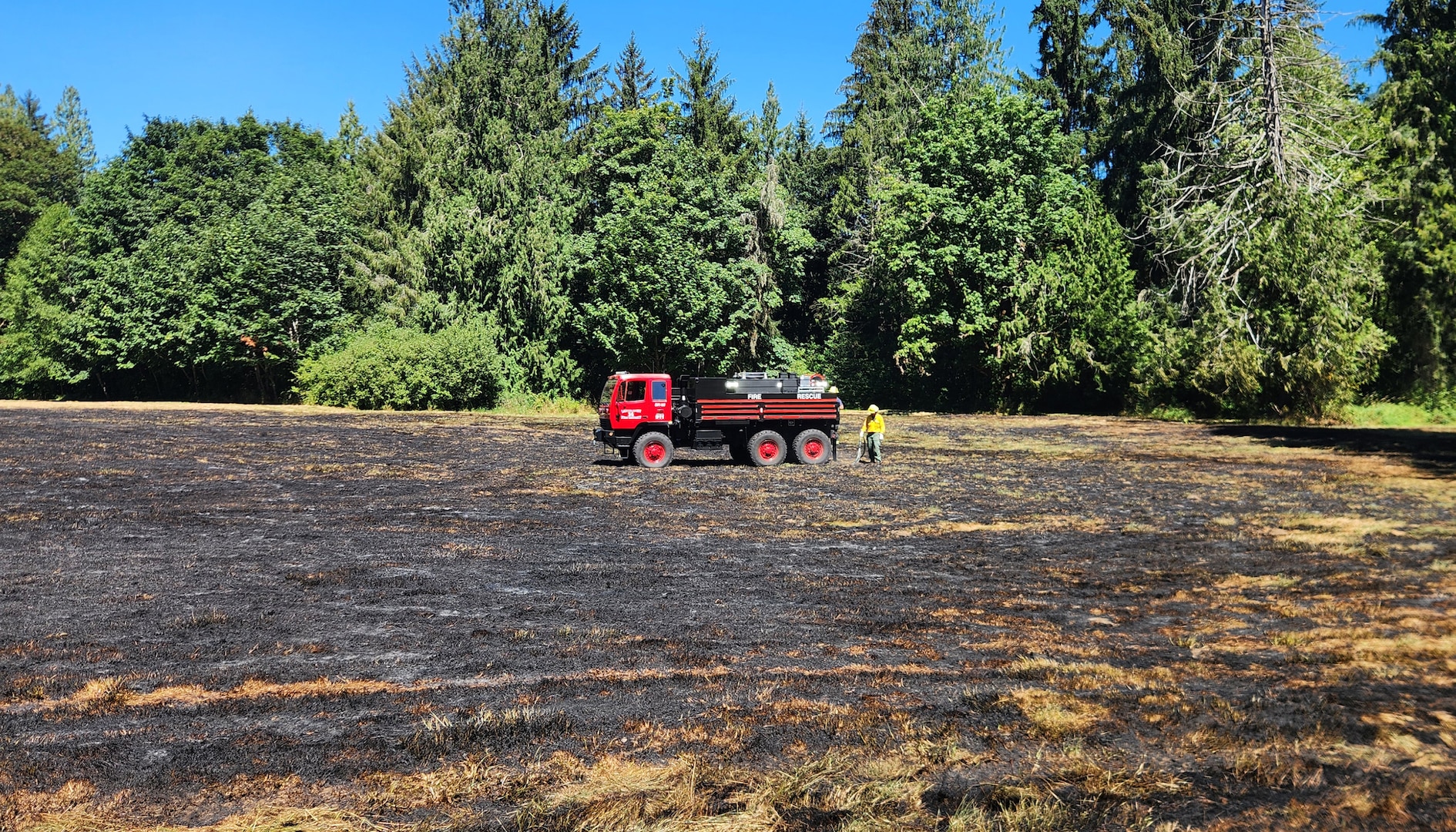 Towards the center of the a burnt grass field a red fire truck and two firefighters can be seen.