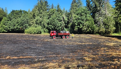 Towards the center of the a burnt grass field a red fire truck and two firefighters can be seen.
