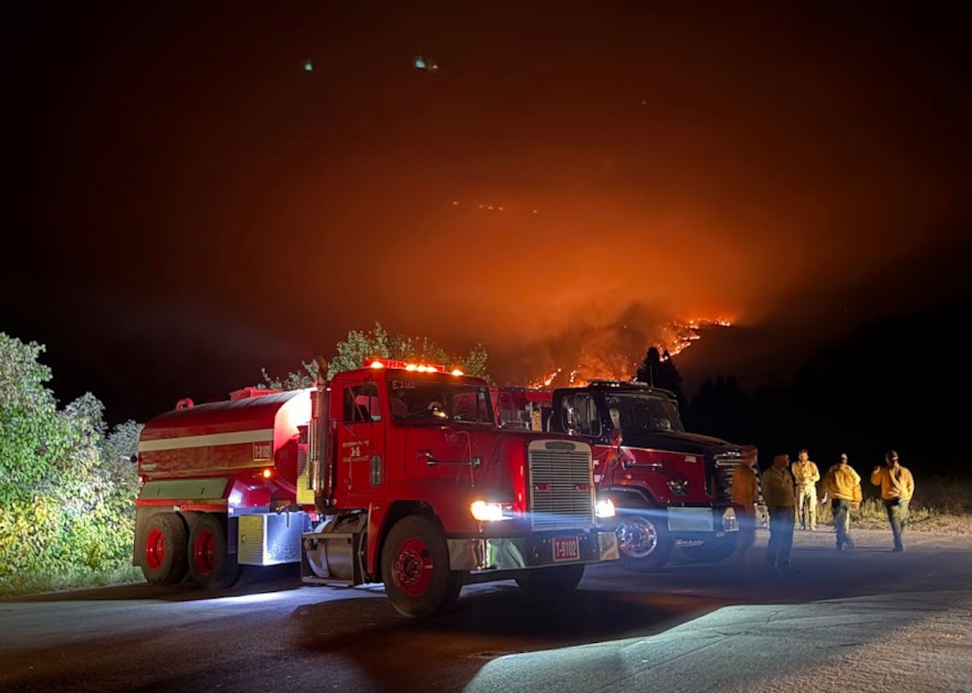 A firetruck sits on a road at night. In the background the glow of a wildfire can be seen. fireman are walking near the truck.
