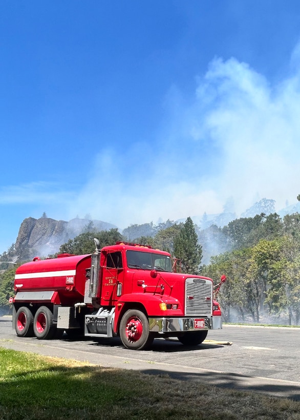 A red fire truck sits on a road next to woods that smoke is rising out of.