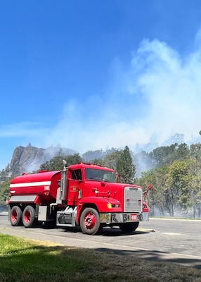 A red fire truck sits on a road next to woods that smoke is rising out of.