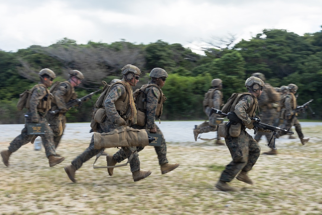 A group of nearly a dozen people wearing camouflage military uniforms and carrying weapons walk briskly past each other with woods behind them.