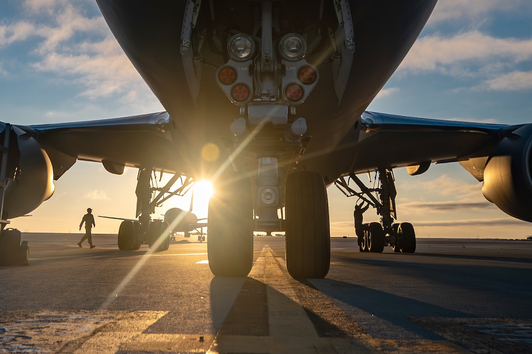 The underside and wheels of a large jet sitting on a runway with a blue sky and clouds above it. People are in the background tending to the vehicle.