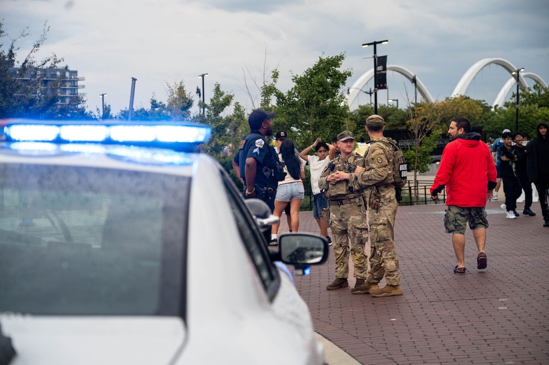 Two people wearing camouflage military uniforms stand on a sidewalk with a police officer, as people walk past. A police car with its blue lights on is visible in the foreground.