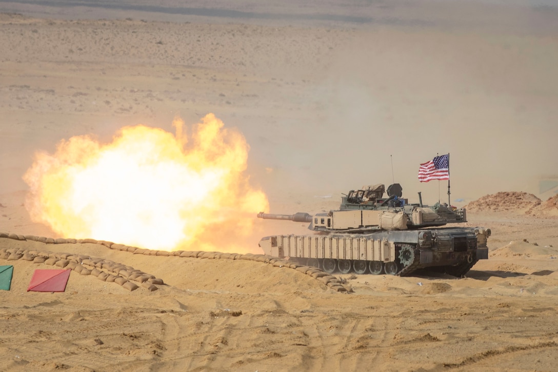 A fireball bursts from a tank gun in a desert environment. There is an American flag mounted on the rear of the tank.