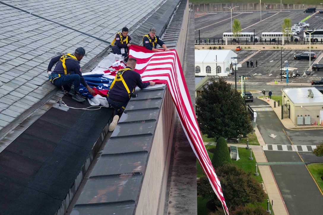 Four people wearing black casual attire and yellow safety harnesses crouch on a building and unfurl an American flag down the side.