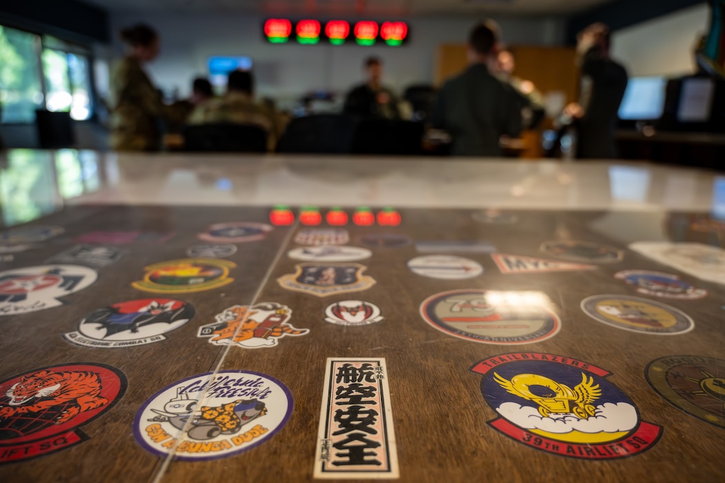 Morale patches are visible in the foreground as Airmen stand in the background in a briefing room