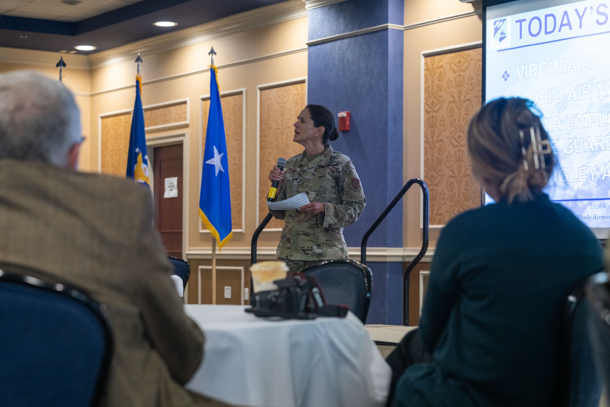 View of BG Jumper addressing the audience, 2 civilians sitting at a table in the foreground