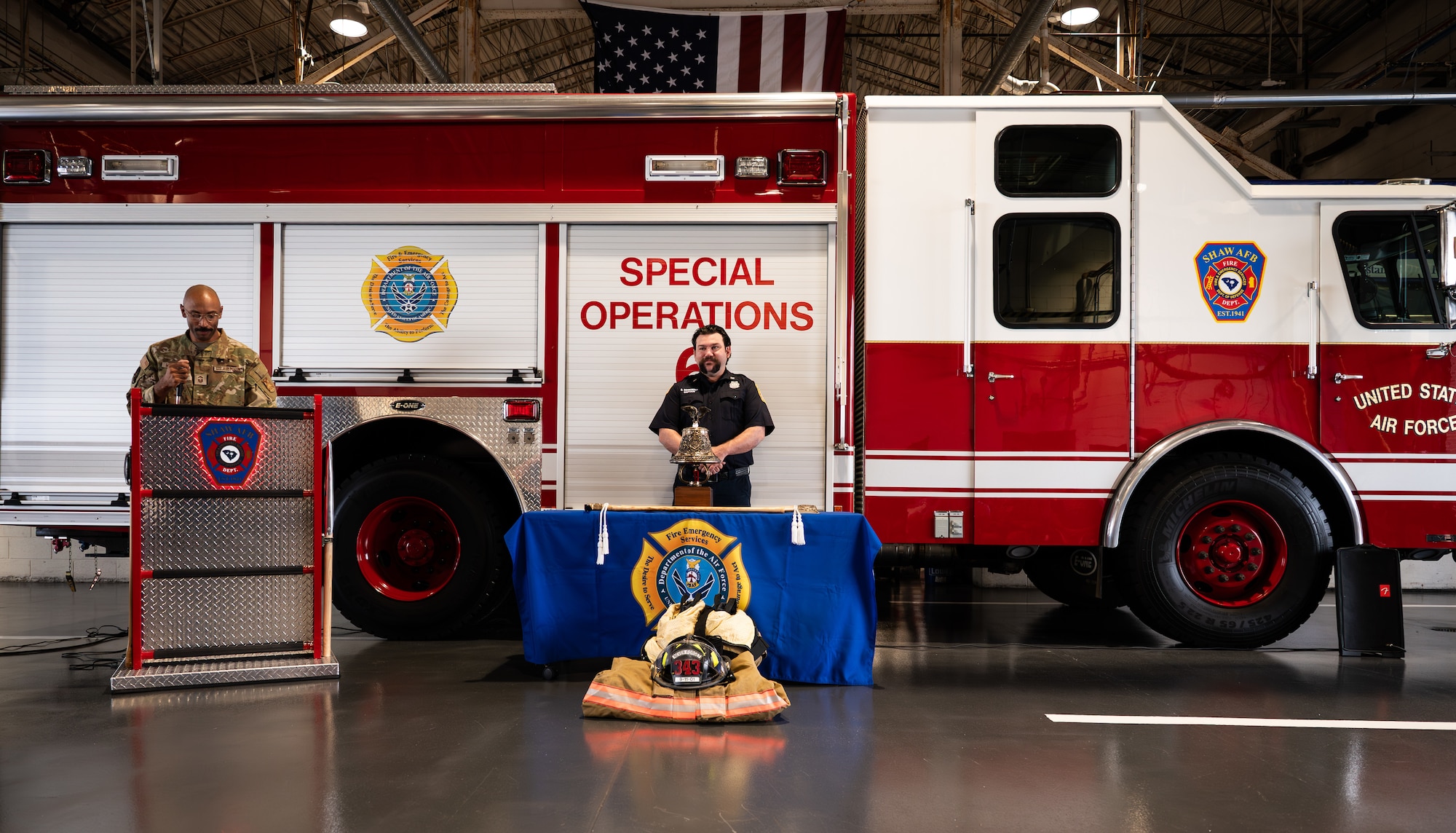 A man in a military uniform and a civilian firefighter stand in front of a fire truck during a speech at a 9/11 memorial service.