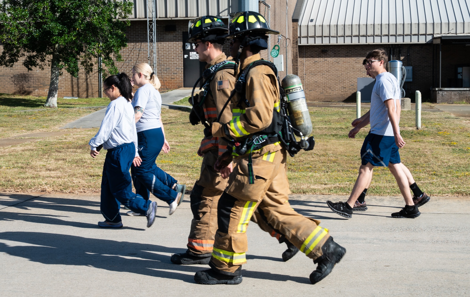 Airmen in PT gear and Airmen in fire fighting gear walk along a road.