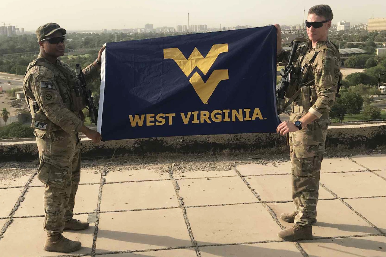 Soldiers in camouflage uniforms hold up a University of West Virginia flag.