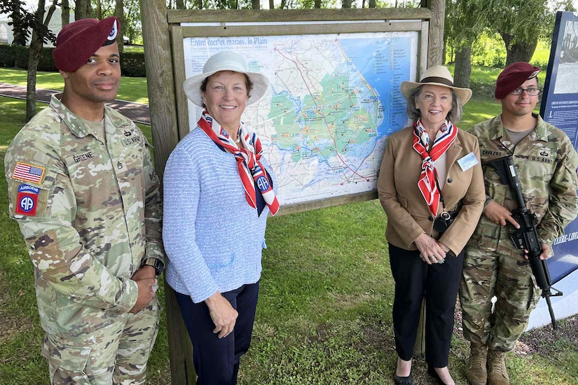 Two soldiers in camouflage uniforms, one holding a rifle, stand next two people dressed in civilian clothing near a map displayed in a grassy area, with trees in the background.