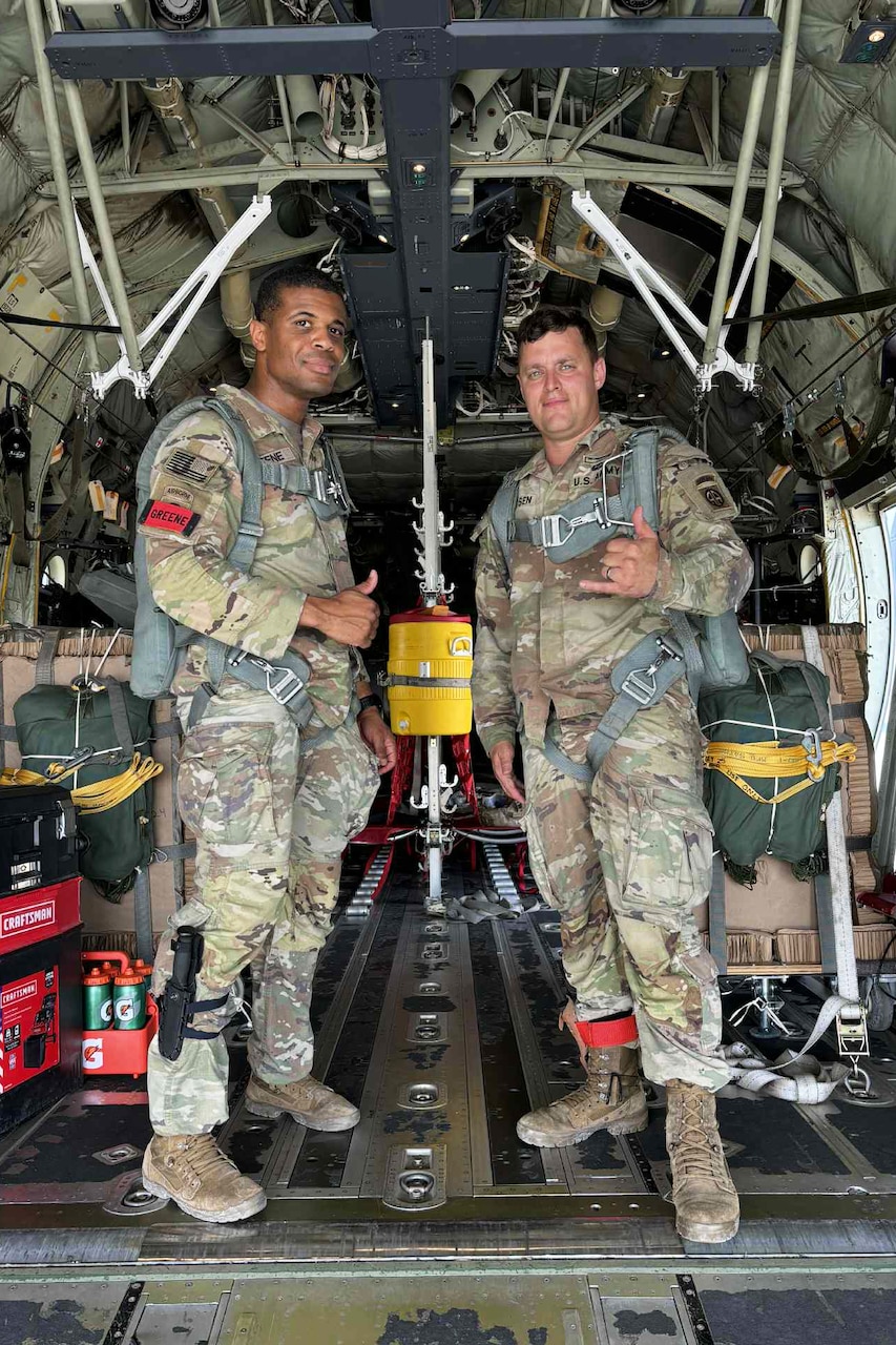 Two soldiers in camouflage uniforms stand in the back of an aircraft, each making a signal with one hand.